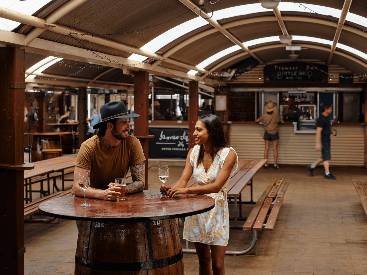 A couple standing at a table drinking a schooner of beer with people standing at the Outback Bottle Shop in the distance