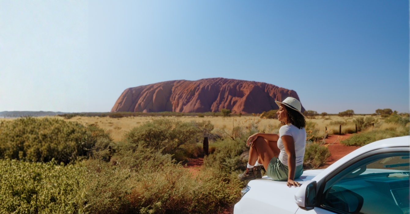 A girl sitting on top of a car with Uluru in the distance