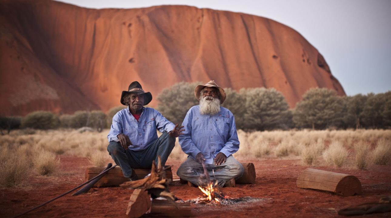 two men sit in front of Uluru by a campfire