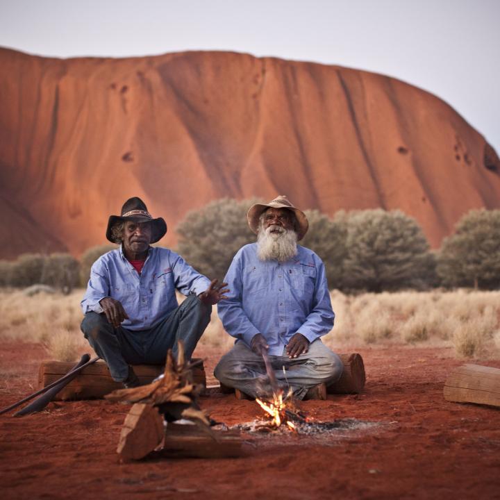 two men sit in front of Uluru by a campfire