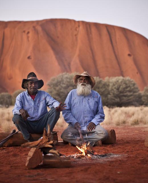 two men sit in front of Uluru by a campfire