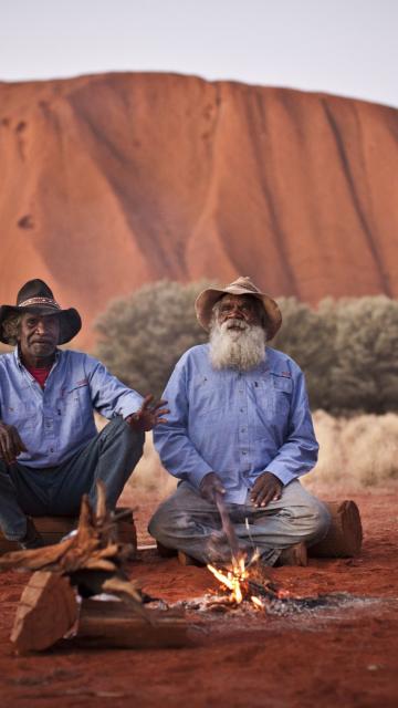 two men sit in front of Uluru by a campfire